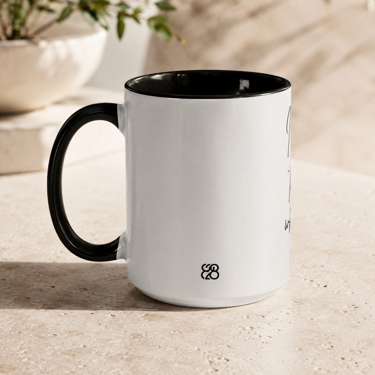 A white ceramic coffee mug with a black handle and black interior, showing the back design with a small 828 Makers Mark, sitting on a light beige stone surface with a blurred plant in the background.