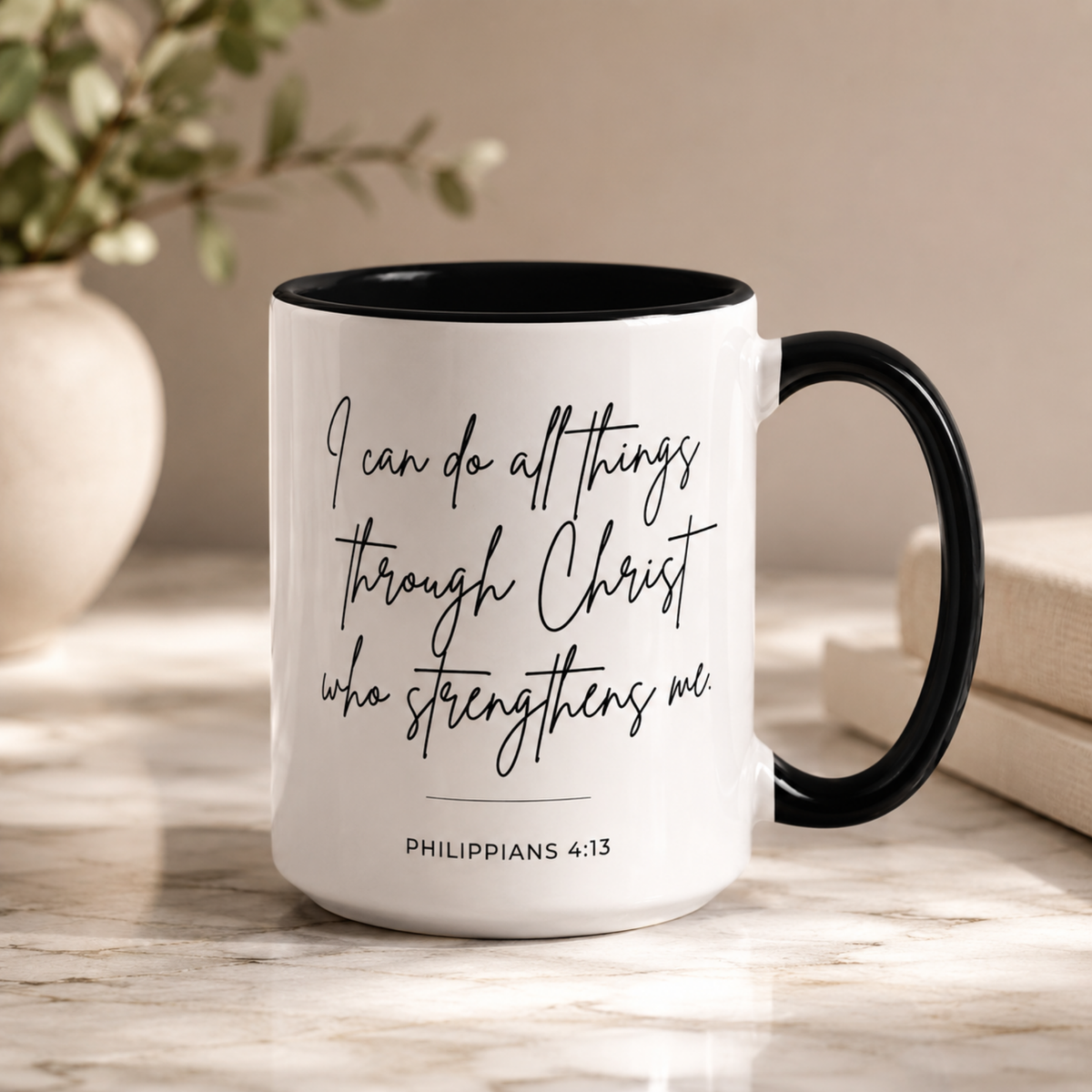 A white ceramic coffee mug with a black handle and black interior, featuring the printed quote 'I can do all things through Christ who strengthens me' and 'PHILIPPIANS 4:13' in black script. The mug sits on a light marble surface with a blurred beige background containing a vase with greenery and books.
