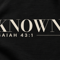 Simulated detail mockup of warm ivory KNOWN. and Isaiah 43:1 transfer artwork on Black fleece; not a manufactured sample photograph.