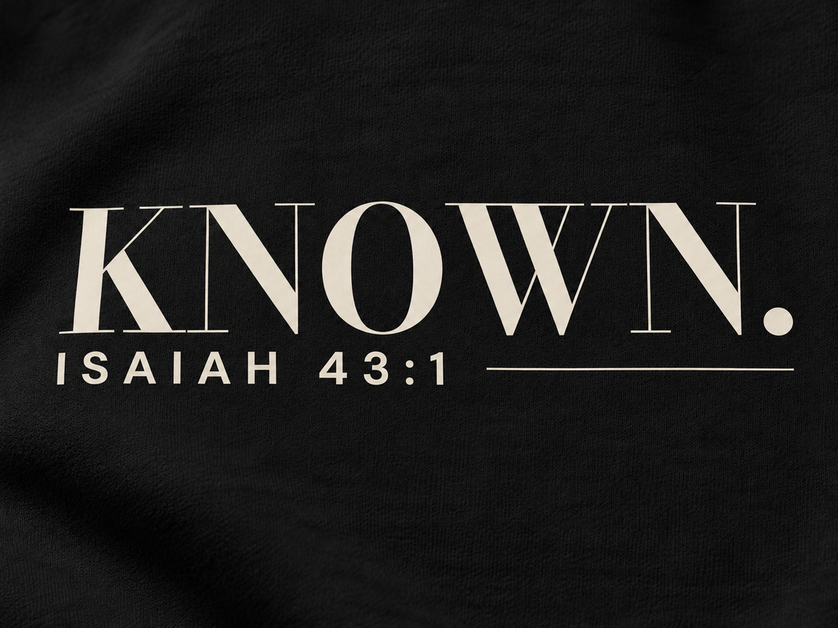 Simulated detail mockup of warm ivory KNOWN. and Isaiah 43:1 transfer artwork on Black fleece; not a manufactured sample photograph.