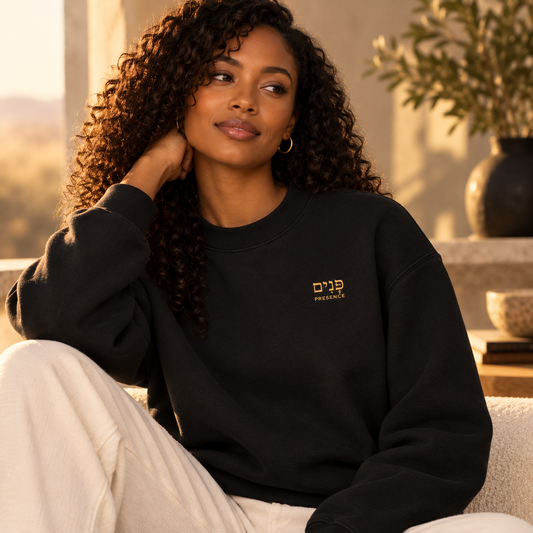 A woman with curly hair wearing a black crewneck sweatshirt with small gold Hebrew embroidery and the word “Presence” on the chest, paired with white pants in a warm indoor setting.