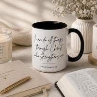 A styled tabletop scene featuring a white ceramic mug with a black handle and black interior printed with the quote 'I can do all things through Christ who strengthens me' from Philippians 4:13. Surrounding props include a candle jar labeled 'FAITH HOPE LOVE,' a notebook with gold text and pen, an open book, and another mug with a scripture quote, all arranged on a light surface with soft natural shadows.