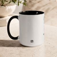 A white ceramic coffee mug with a black handle and black interior, showing the back design with a small 828 Makers Mark, sitting on a light beige stone surface with a blurred plant in the background.