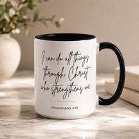 A white ceramic coffee mug with a black handle and black interior, featuring the printed quote 'I can do all things through Christ who strengthens me' and 'PHILIPPIANS 4:13' in black script. The mug sits on a light marble surface with a blurred beige background containing a vase with greenery and books.